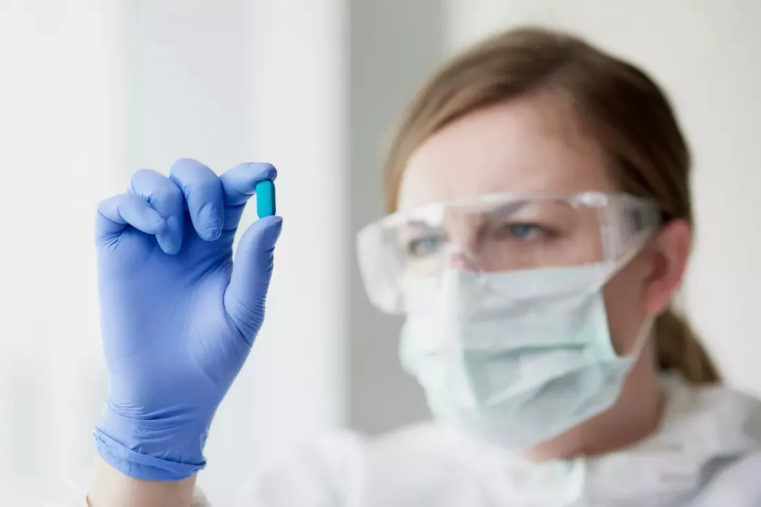 Une laborantine avec lunettes de protection et masque observe une gelule bleue.